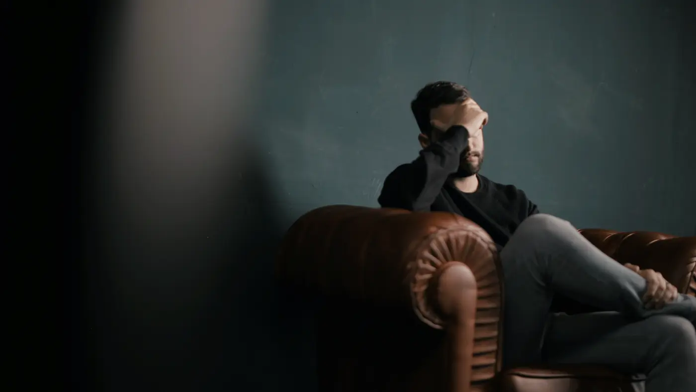 Man sitting in a leather armchair in a dark room with his head in his hand