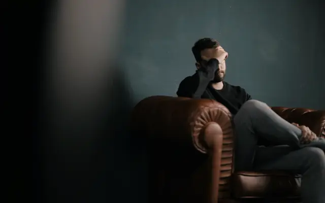 Man sitting in a leather armchair in a dark room with his head in his hand