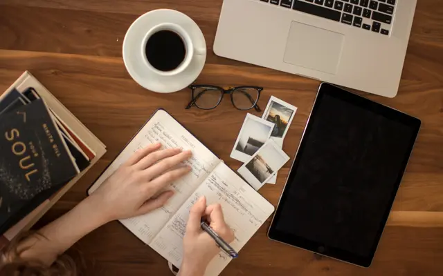 Overhead view of a hand writing in a notebook beside a laptop and coffee