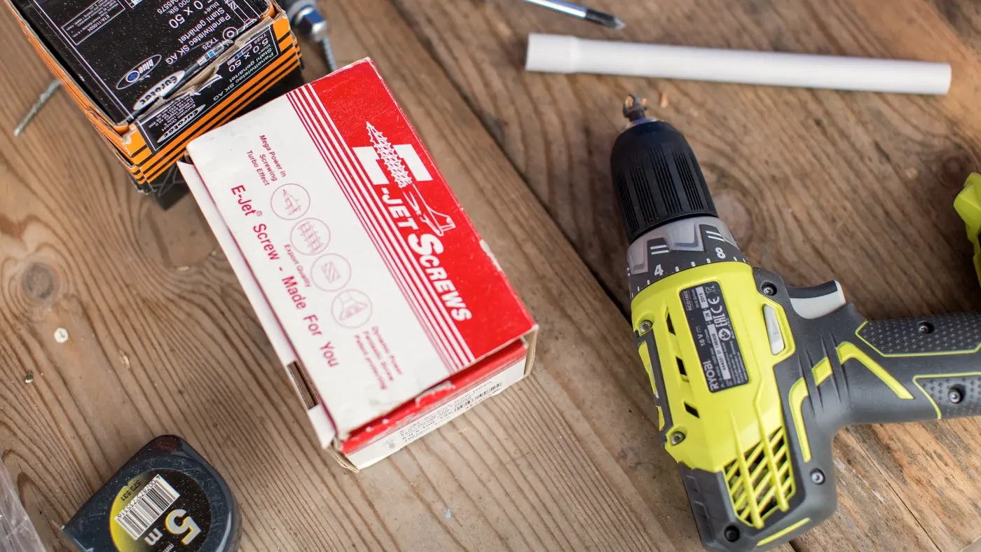 Cordless drill, tape measure and a box of fasteners on a wooden workbench