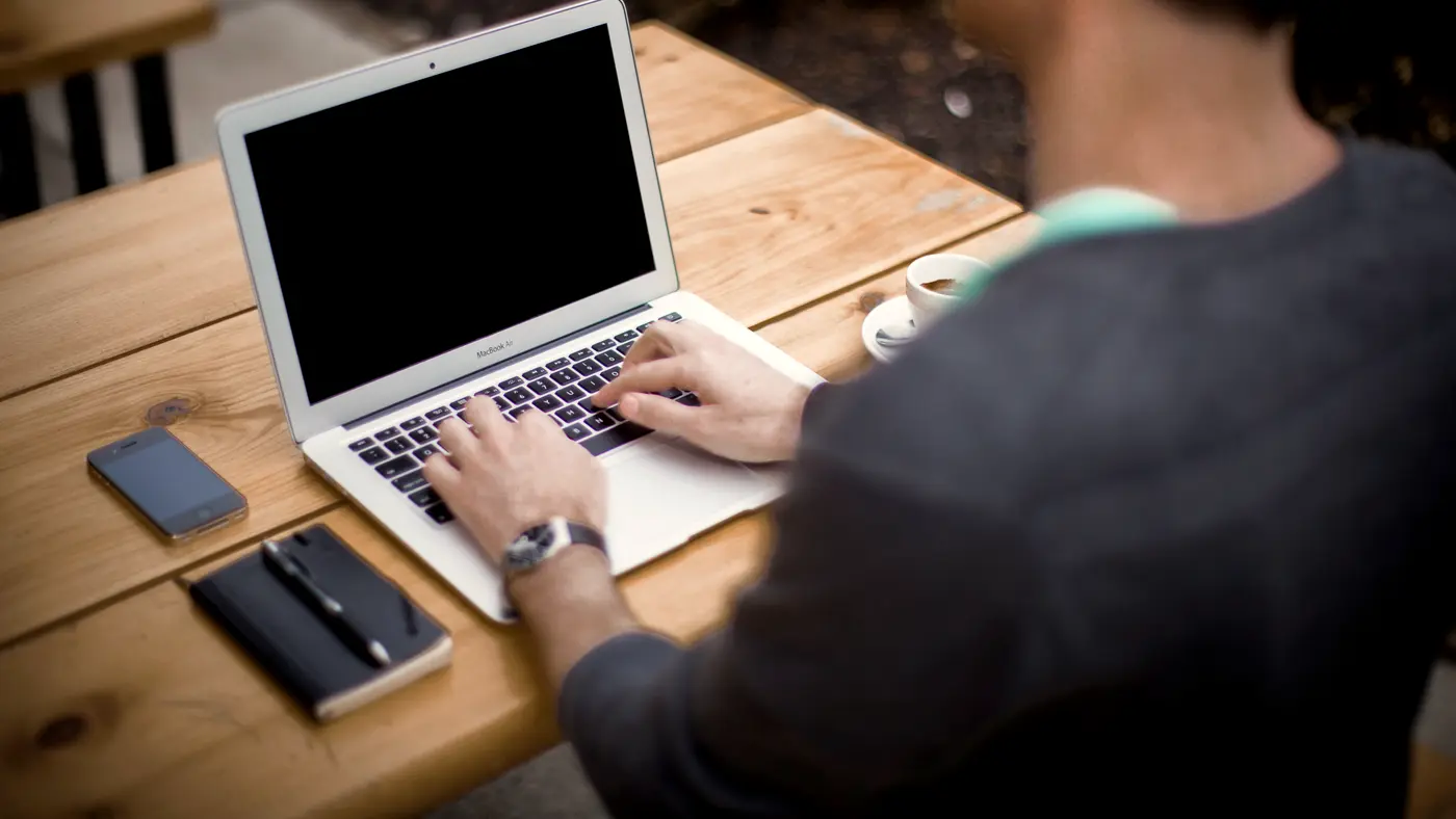 Man typing on a laptop at a wooden table with a notebook and phone