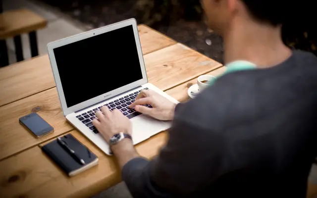 Man typing on a laptop at a wooden table with a notebook and phone